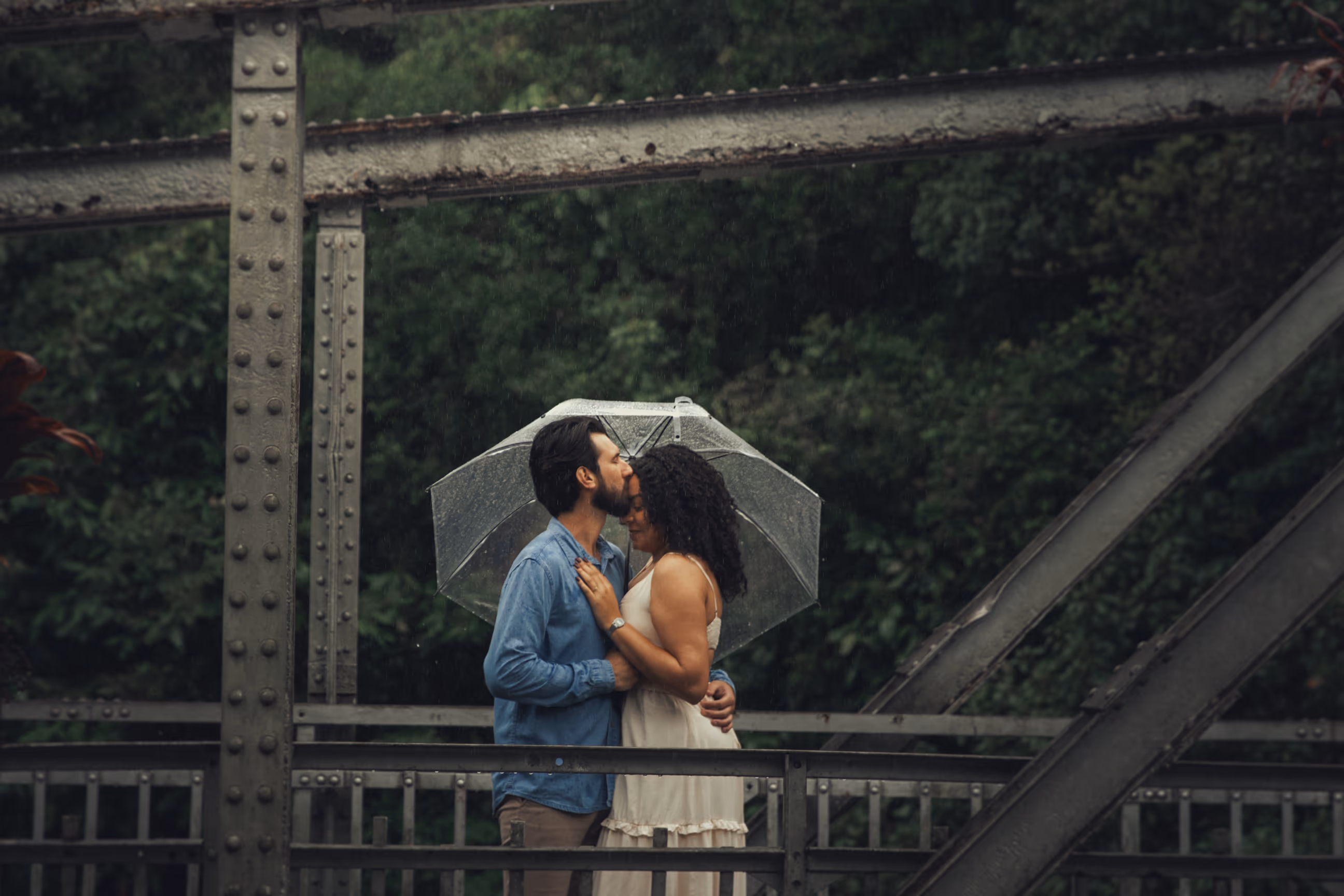 Casal se beijando sob guarda-chuva transparente numa ponte de ferro