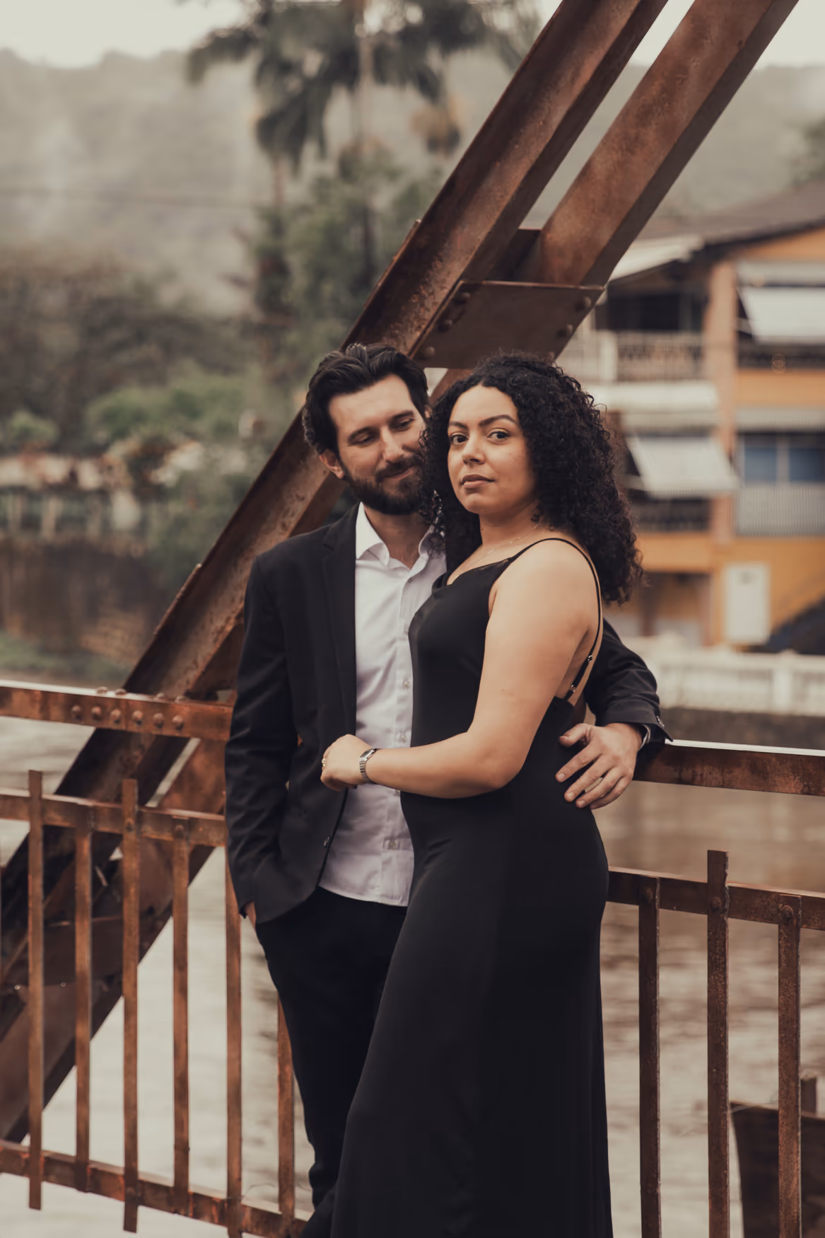 Casal abraçado em uma ponte de ferro com a cidade ao fundo